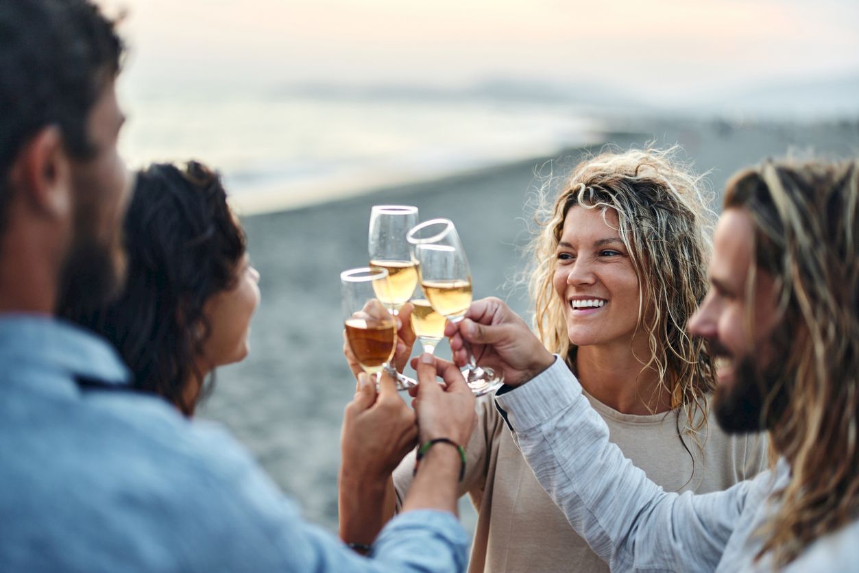 Friends clink champagne flutes outdoors by the ocean at sunset, laughing and toasting.