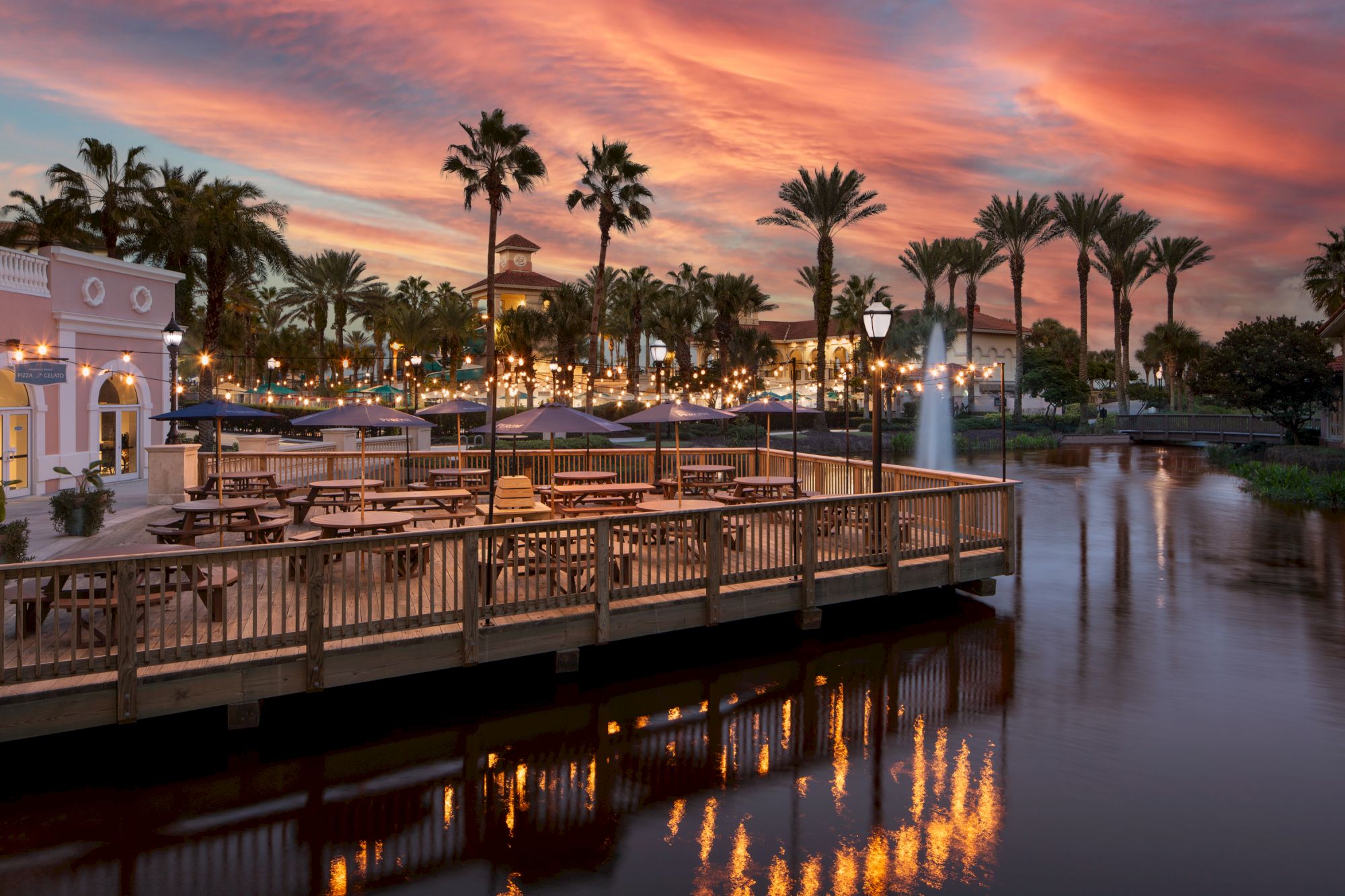 A scenic view of a waterfront café with palm trees, tables, and a sunset sky reflecting on the water, creating a tranquil atmosphere.
