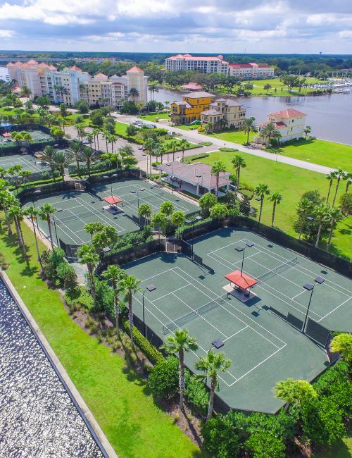 Aerial view of several tennis courts surrounded by palm trees near a waterfront, with buildings and a small marina in the background.