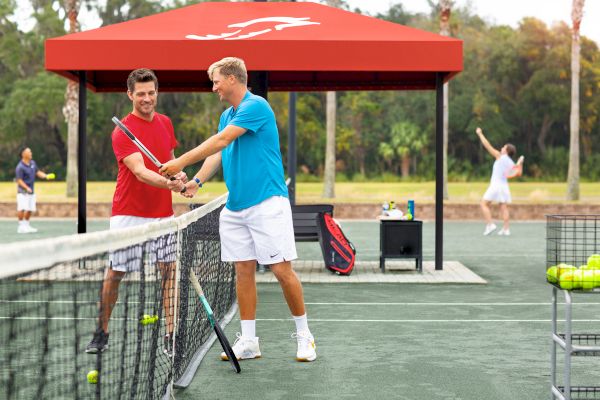 Two people on a tennis court, with one guiding the other on racket technique. Others are playing in the background under a red canopy.