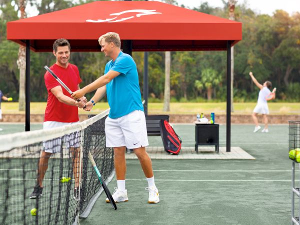Two people on a tennis court, with one guiding the other on racket technique. Others are playing in the background under a red canopy.