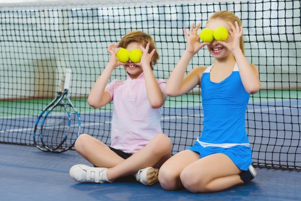 Two children sit on a tennis court, playfully holding tennis balls over their eyes with rackets nearby by the net.