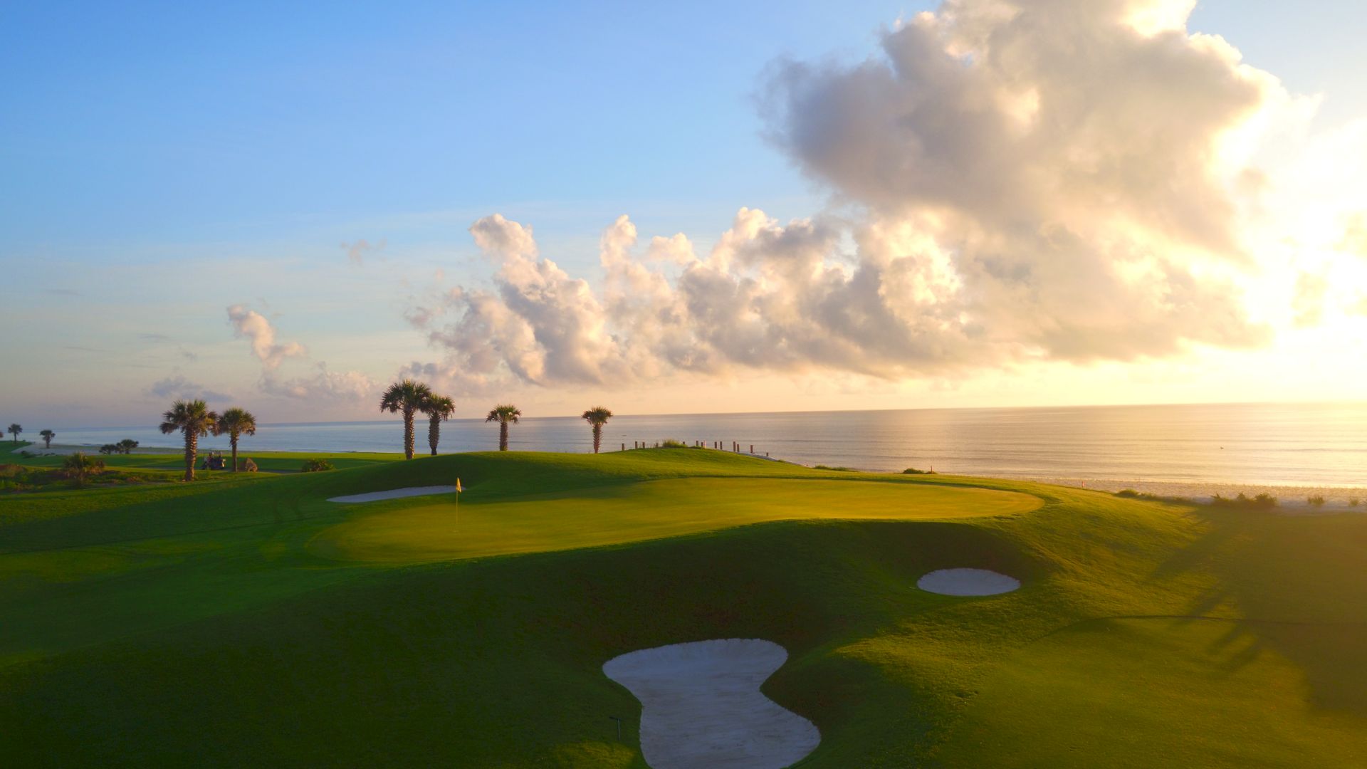 A golf course by the sea during sunset, with palm trees, sand traps, and a vast sky covered in fluffy clouds.