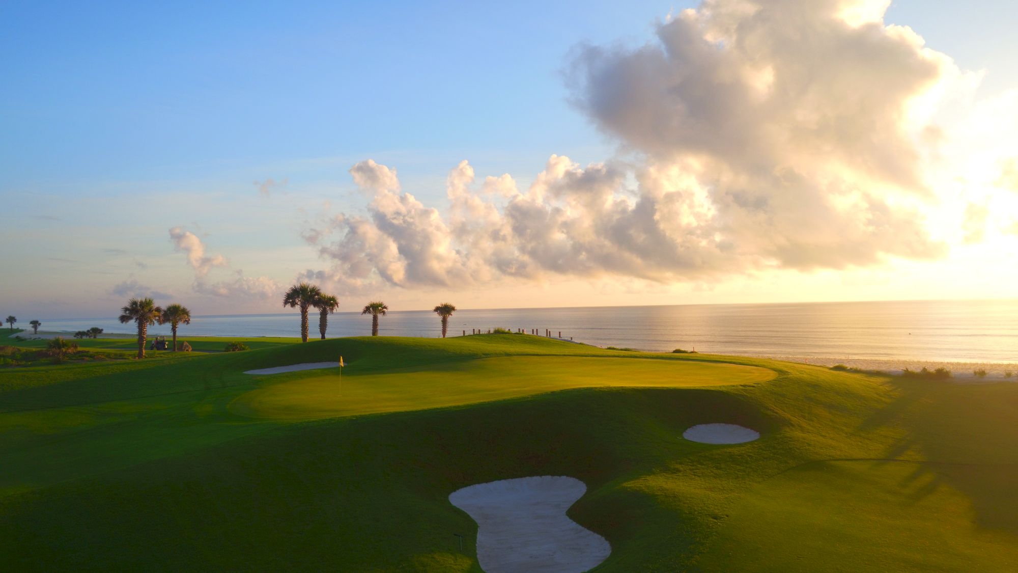 A golf course by the sea during sunset, with palm trees, sand traps, and a vast sky covered in fluffy clouds.