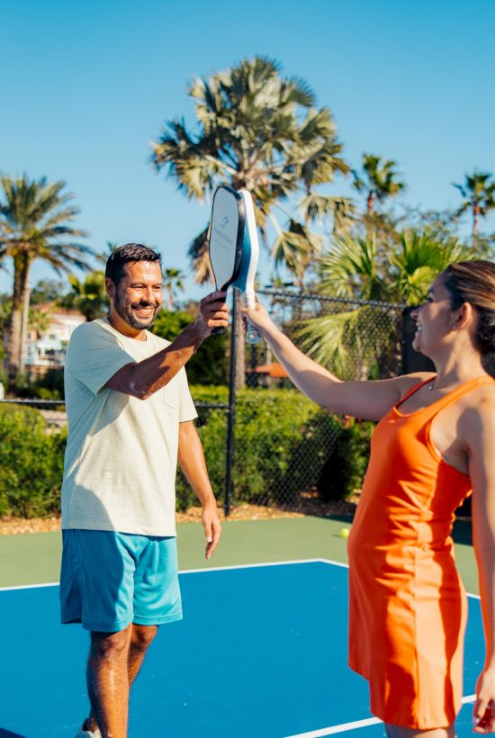 Two people on a sunny tennis court high-five with their rackets, surrounded by palm trees and a clear blue sky.