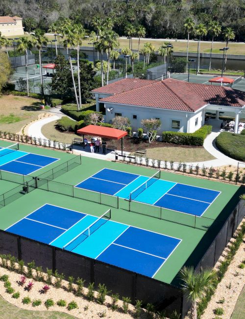 An aerial view of multiple pickleball courts next to a clubhouse and surrounded by trees and landscaped areas.