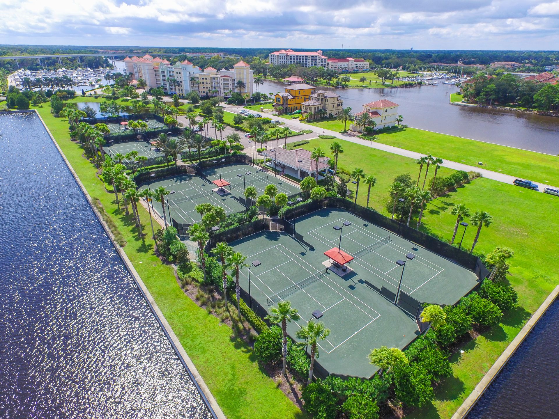 This aerial view shows tennis courts surrounded by trees, near water, with buildings and a marina visible in the background.
