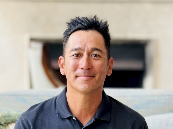 A person in a navy polo shirt is sitting on a sofa, smiling at the camera, with a blurred background behind them.