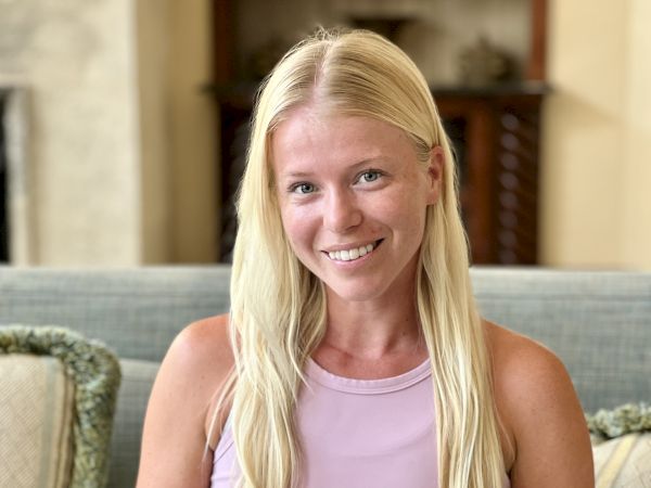 A person with long blonde hair is sitting on a sofa, wearing a pink top and smiling at the camera.