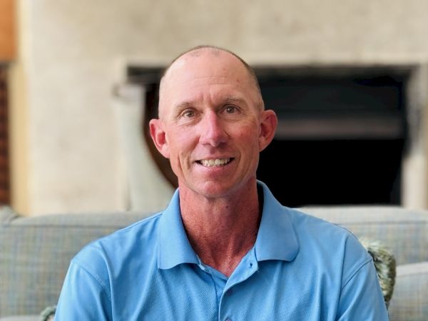 A person wearing a blue polo shirt is sitting on a sofa with a neutral background.