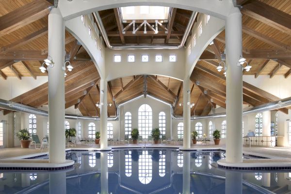A spacious indoor swimming pool with vaulted wooden ceilings, elegant columns, and large windows letting in natural light.