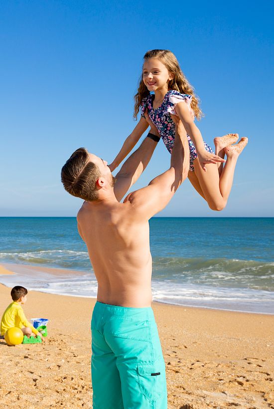 A man joyfully lifts a child on a sunny beach with blue umbrellas and people relaxing in the background by the ocean.