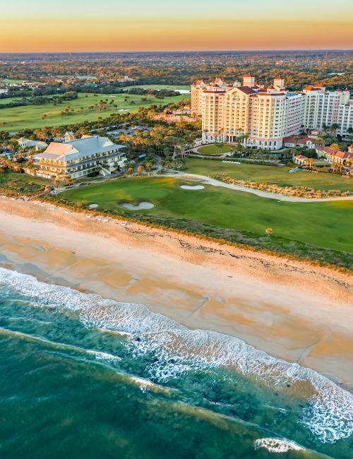 Aerial view of a beachfront resort with buildings, golf course, and ocean waves, set against a clear sky during sunset.
