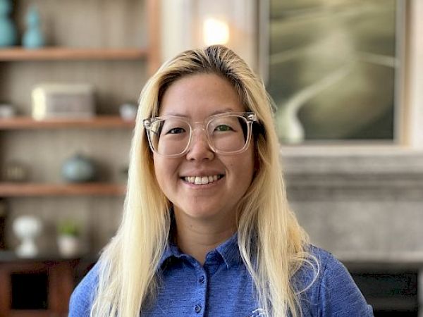 A person with long hair and glasses in a blue polo shirt stands indoors, smiling. They're in a room with decorative items on shelves.