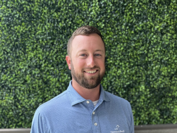 A man in a blue polo shirt stands smiling against a backdrop of green foliage and a wooden floor, with a logo on his shirt.