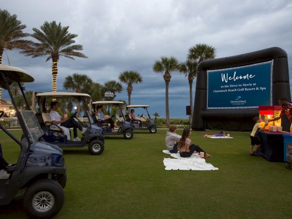 An outdoor movie event with people in golf carts and on blankets, facing a large screen on a lawn with palm trees and a concession stand.