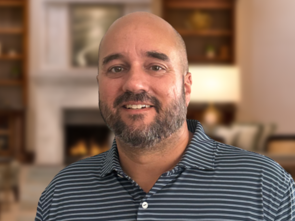 A man in a striped polo shirt stands smiling in a cozy room with a fireplace and bookshelves in the background.