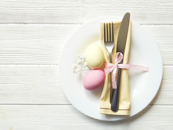 A white plate holds pastel eggs, cutlery wrapped in a napkin with a pink ribbon, and small flowers, all on a wooden surface.