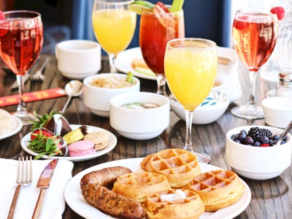 A brunch table with waffles, sausage, fruit, macarons, and assorted drinks, including orange juice and red cocktails.