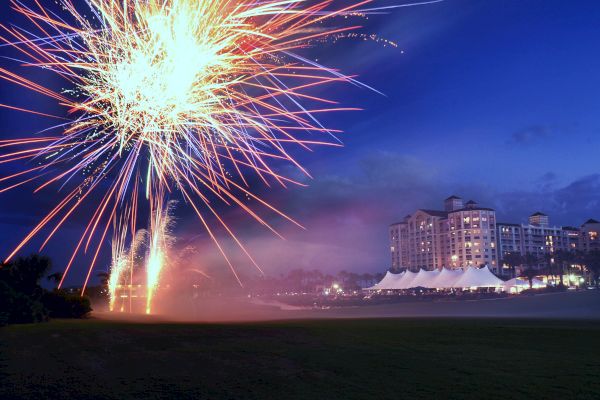 A vibrant fireworks display illuminates the night sky near a large, well-lit building with tents and palm trees.