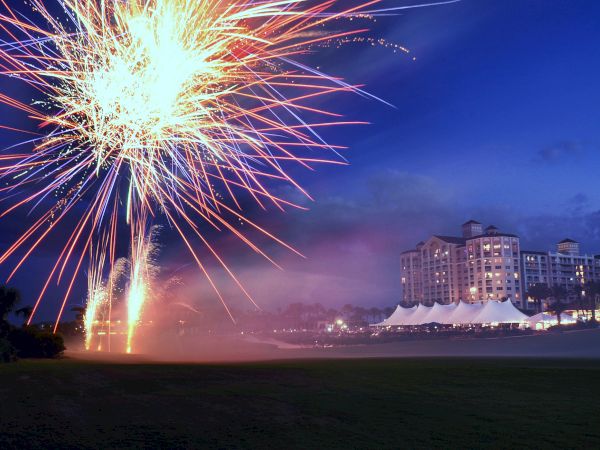 A vibrant fireworks display illuminates the night sky near a large, well-lit building with tents and palm trees.