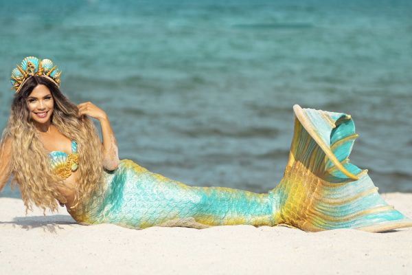 A person dressed as a mermaid with long hair and a detailed tail, relaxing on the beach near the ocean.