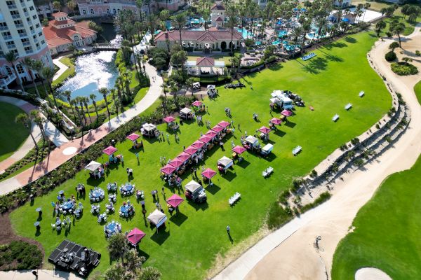 An outdoor event with tables, chairs, and tents on a green field near a resort and pool, with people attending and carts nearby.