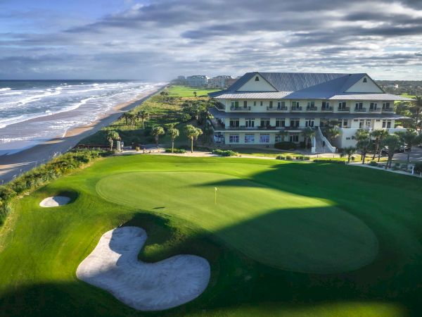 A coastal golf course features a putting green, bunker, and a clubhouse, set against ocean waves and a cloudy sky.