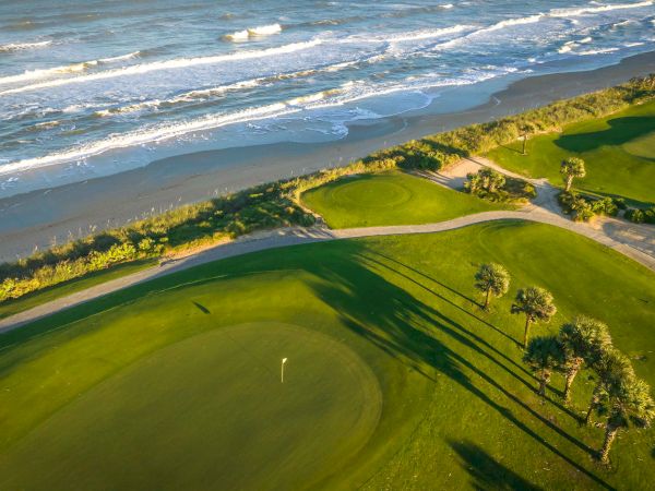 Aerial view of a coastal golf course featuring lush green fairways, palm trees, and ocean waves along a sandy shore.