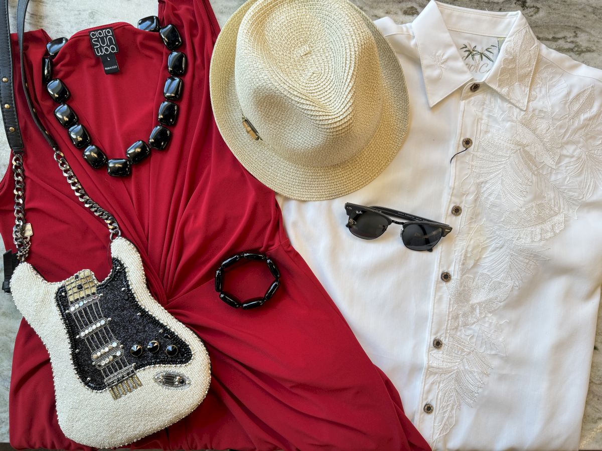 A red dress, white shirt, straw hat, guitar-shaped purse, black necklace, bangle, and sunglasses are artfully arranged on a surface.