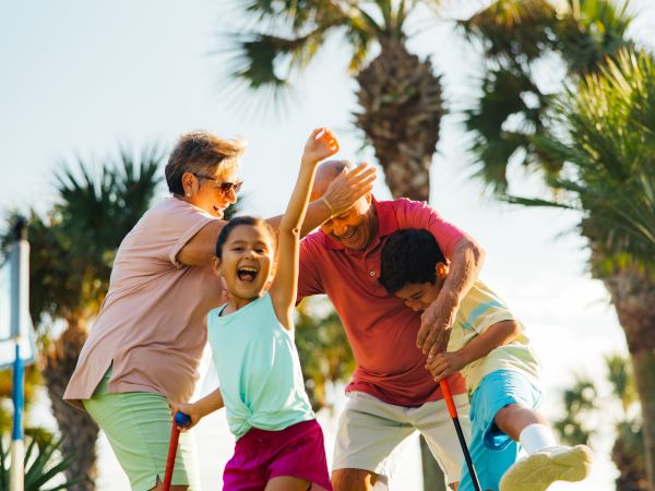 A joyful family plays mini-golf outdoors, with palm trees in the background and sunlight creating a fun and lively atmosphere.