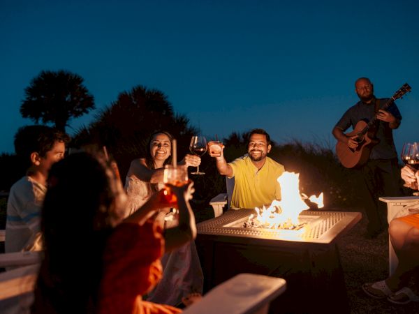 A group of people is gathered around a fire pit, toasting with drinks, while someone plays guitar in the background during the evening.