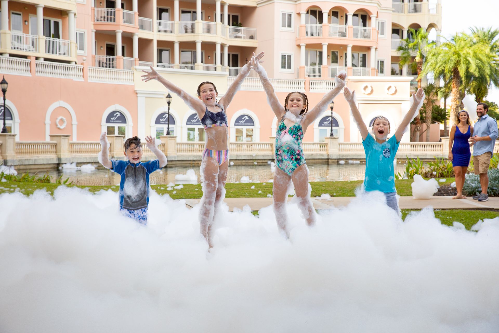 Children joyfully playing in foam outside a large building, with two adults in the background observing them.