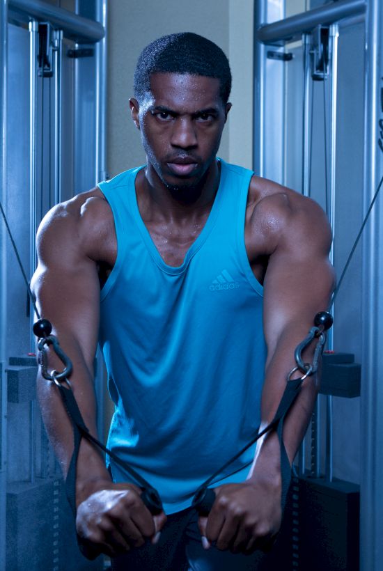 A man in a blue tank top is exercising on a cable machine in a gym setting with dramatic lighting and mist.