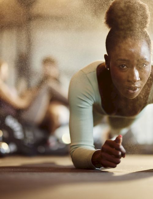 A person in workout attire doing a plank exercise in a gym, with a water bottle to the side, and others in the background.