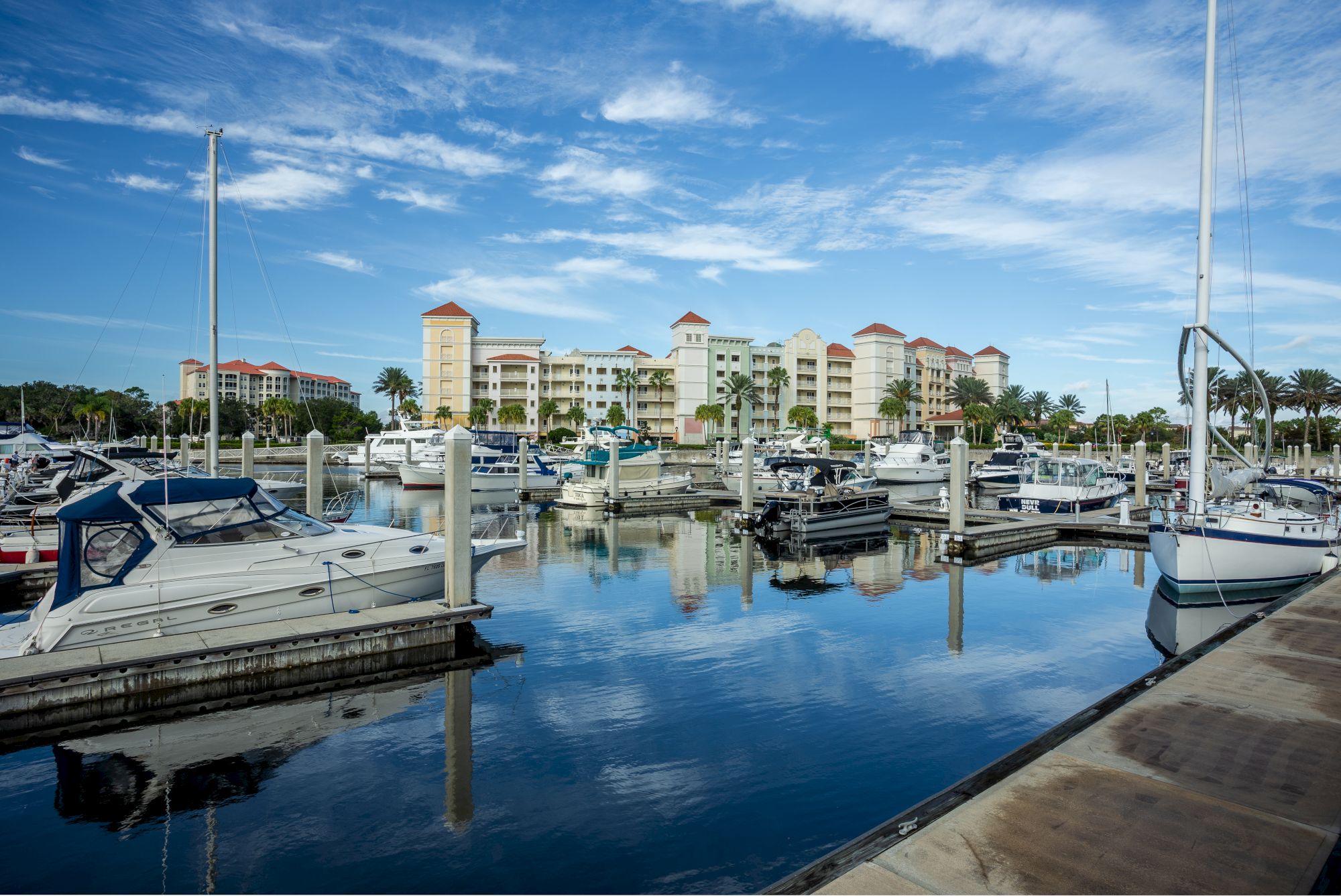 A marina with boats docked, calm water reflecting the sky and surrounding buildings, under a partly cloudy blue sky.