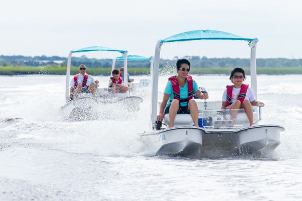 People are enjoying a ride on small motorized boats on a body of water, wearing life jackets and surrounded by lush greenery.