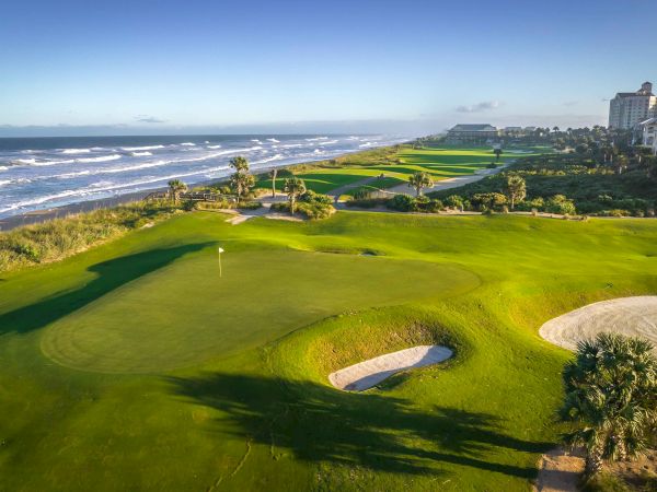 A golf course by the ocean, featuring lush greens and sand traps, with buildings in the distance under a clear blue sky.