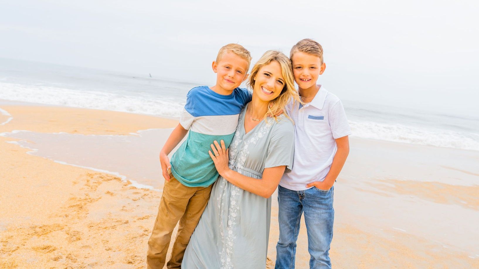 A woman and two boys smile while standing on a beach, with waves in the background.