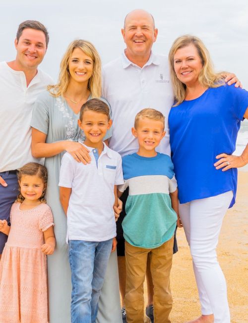 A family of seven stands on a sandy beach, smiling, with the ocean in the background under a cloudy sky.