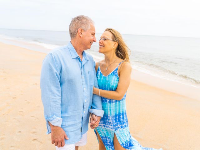 A couple is standing on a beach, smiling at each other while holding hands.