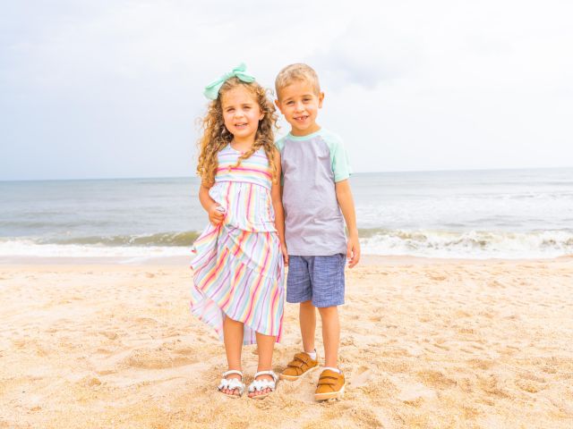 Two children stand on a sandy beach near the ocean, smiling. One wears a colorful dress, the other a T-shirt and shorts. Waves in the background.