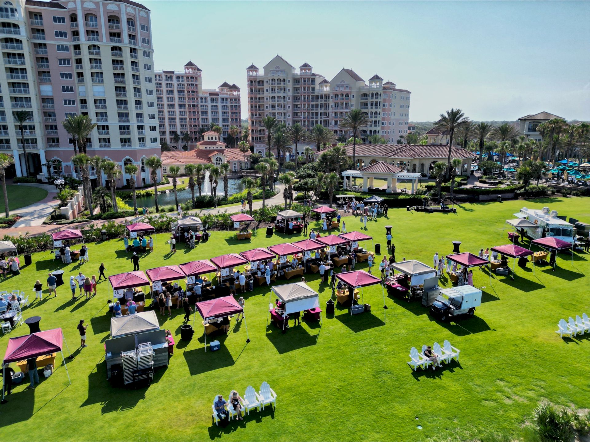 Aerial view of an outdoor event with stalls and people on a grassy area, surrounded by tall buildings and palm trees in the background.