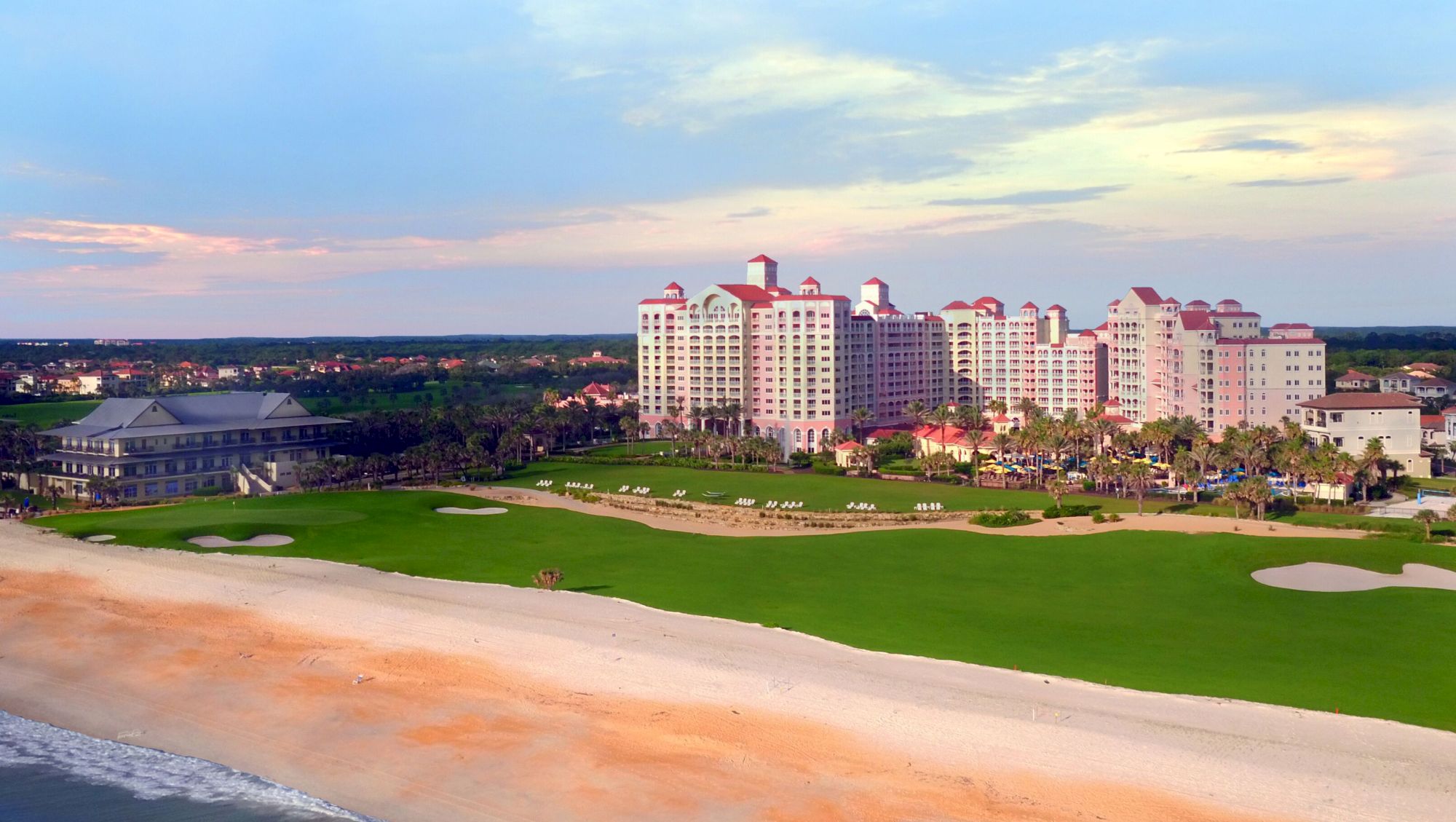 The image shows a large beachfront resort with multiple buildings, surrounded by a golf course and a sandy coastline.