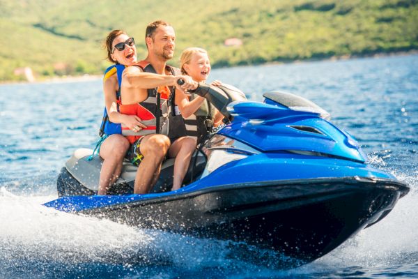 A family of three is enjoying a ride on a jet ski on the water, with scenic greenery in the background.
