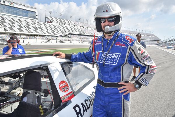 A person in a racing suit and helmet stands by a race car on a track. Another person with a headset is in the background.