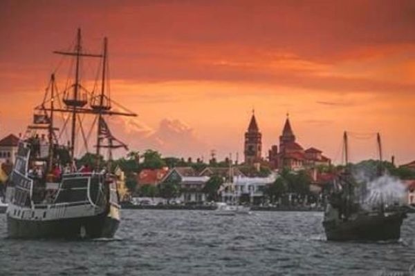 Ships sail in a harbor with a historic town and church steeples in the background, under a dramatic orange sky.