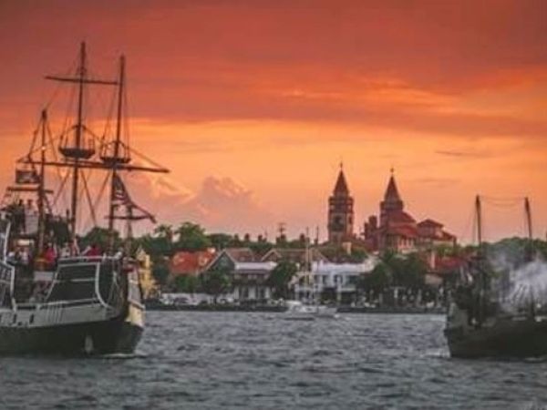 Ships sail in a harbor with a historic town and church steeples in the background, under a dramatic orange sky.