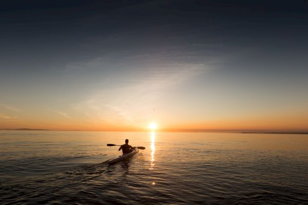 A person is kayaking on calm waters toward a sunset under a vast sky, creating peaceful reflections on the ocean surface.