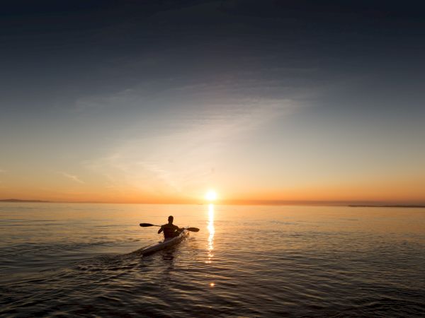 A person is kayaking on calm waters toward a sunset under a vast sky, creating peaceful reflections on the ocean surface.